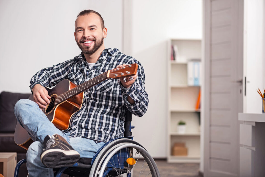 Man wearing plaid shirt who uses a wheelchair smiles as he plays guitar